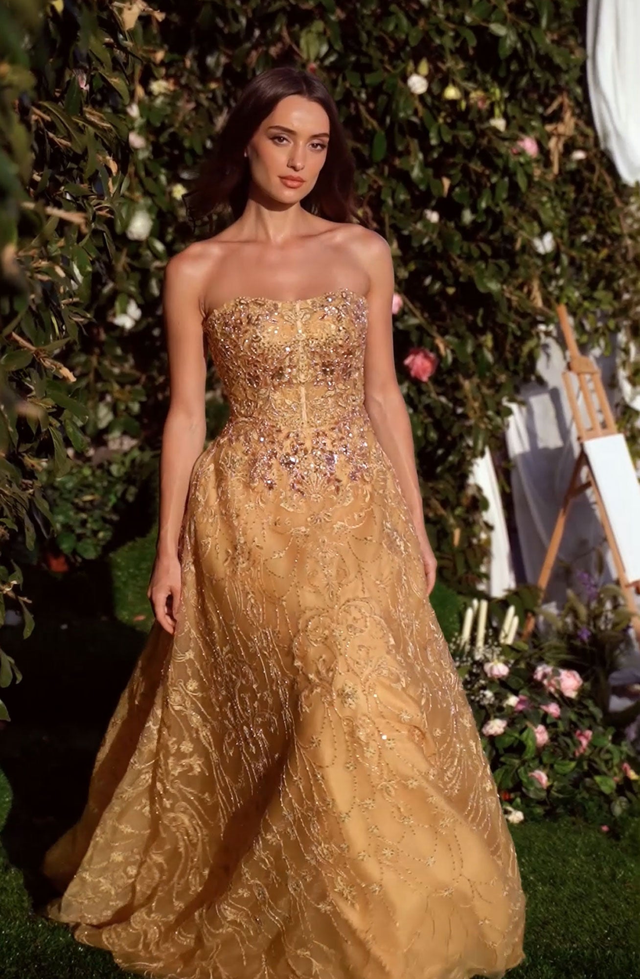 A woman wearing the Andrea & Leo MA104 long sleeve long formal dress stands outdoors near greenery and flowers, with an easel and candles in the background.